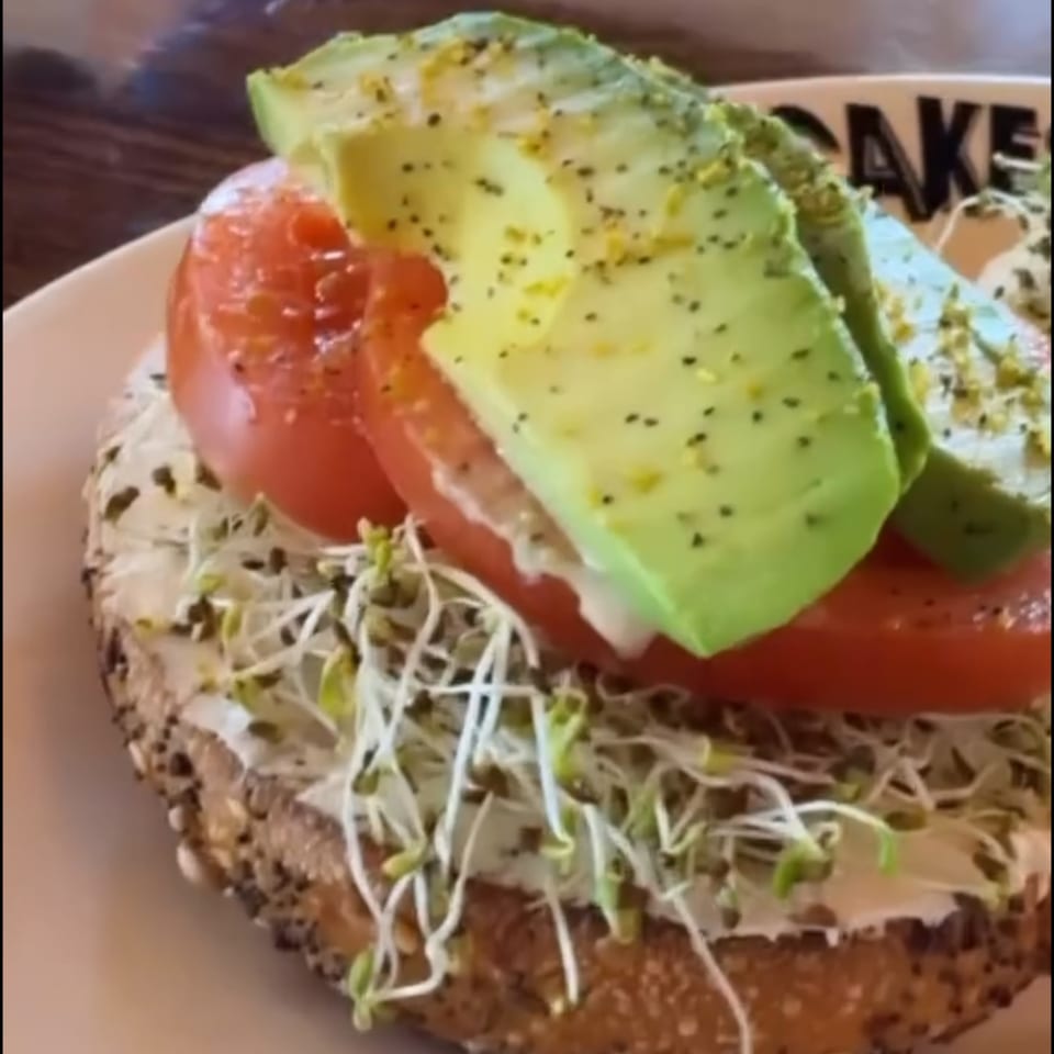 Close-up of a bagel with cream cheese, sprouts, tomato slices, and avocado, on a white plate.
