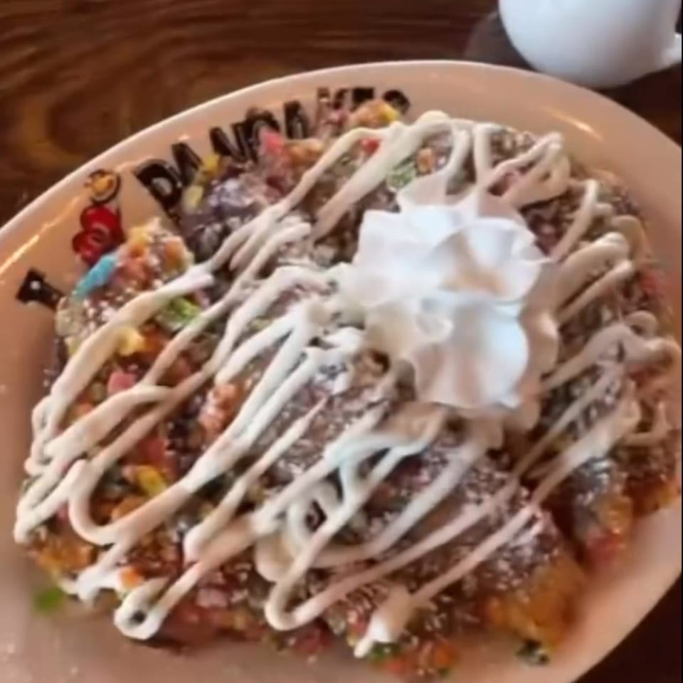 A large pancake from I Heart Pancakes, topped with whipped cream, icing, and colorful cereal.