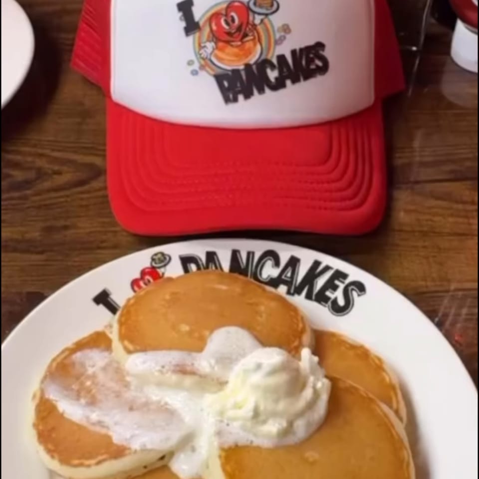 Fluffy pancakes with whipped cream and syrup on a plate, with an "I Heart Pancakes" hat.