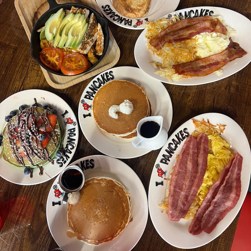 A table laden with various breakfast dishes from I Heart Pancakes, including pancakes, bacon, and eggs.